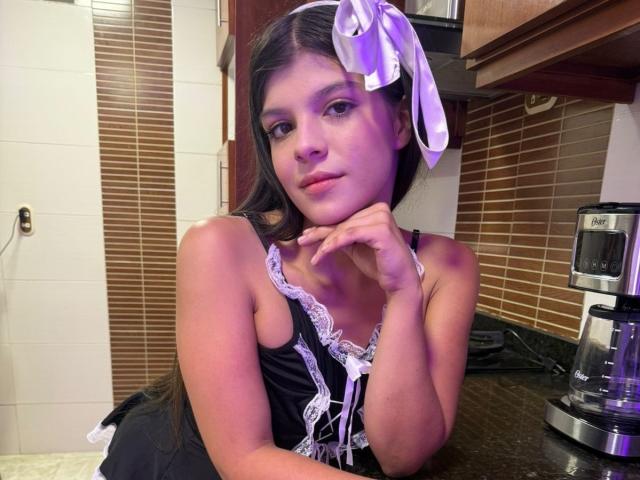 RilleyFoyx with purple eyes, wearing a black and white lace maid outfit, has a purple satin bow in her long brown hair, leaning on a kitchen counter.