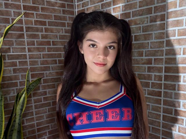RilleyFoyx with long dark hair in pigtails, wearing a blue and red cheerleader top, showing small tits, standing in front of a brick wall with a plant on the left.