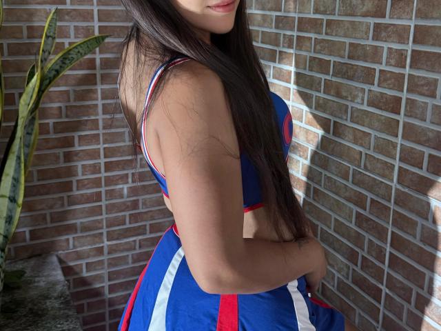 RilleyFoyx with long dark hair, wearing a blue, red, and white dress, showing her ass, against a brick wall, with a snake plant on the left.
