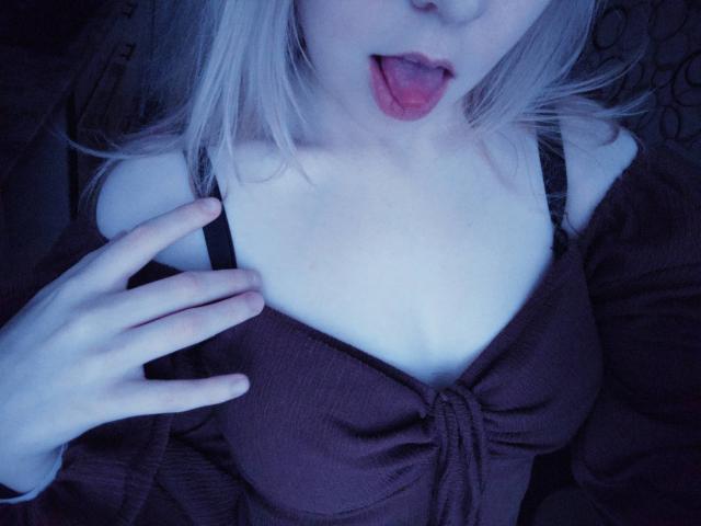 TiffaniMacandog with pale skin, blonde hair, and tongue out, holding her black-strapped brown top, exposing her tits, in a blue-lit selfie. Her hand is on her chest.