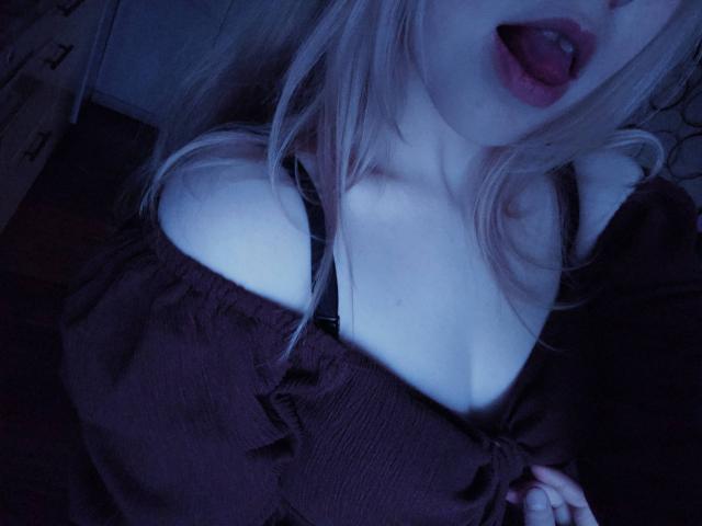 TiffaniMacandog's tongue is out, her lips are parted, showing red lipstick. She's wearing a dark off-shoulder top that reveals her black bra and cleavage. Image is blue-tinted.