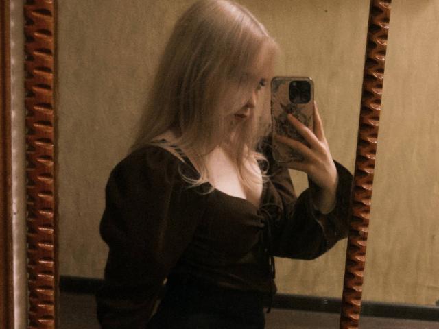 TiffaniMacandog with long blonde hair, wearing a brown top, taking a mirror selfie with her phone, showing her tits and side profile, in a dimly lit room.