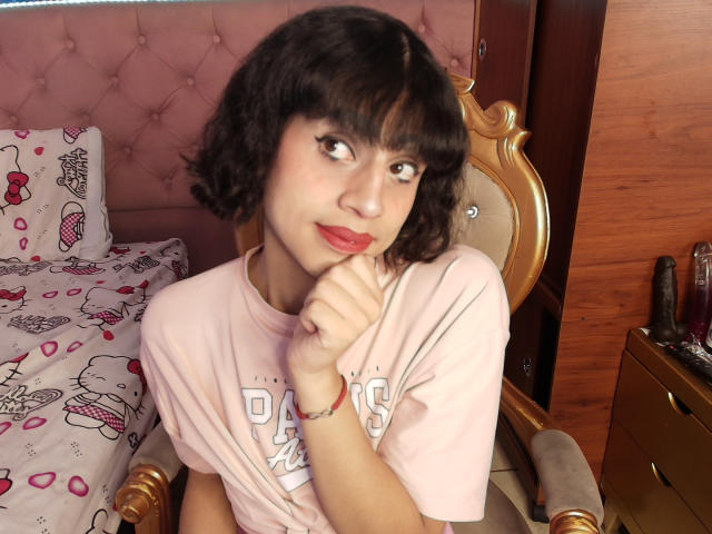 EimyFlorian has short black curly hair, light skin, and red lipstick. She's wearing a pink shirt, sitting on a wooden chair with her finger on her chin.