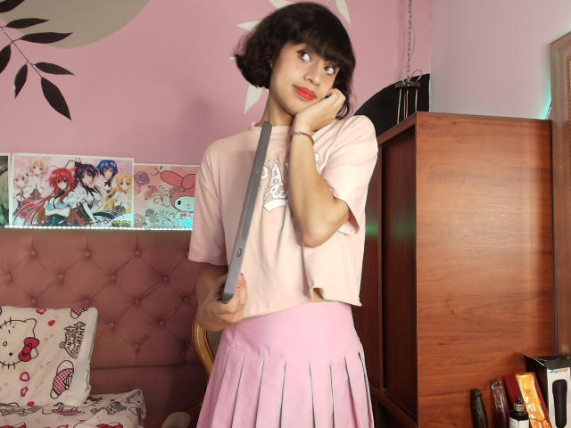 EimyFlorian with short black hair, pink top, and pink pleated skirt, holding a knife to her cheek, looking up with red lipstick, in a pink, anime-themed room.