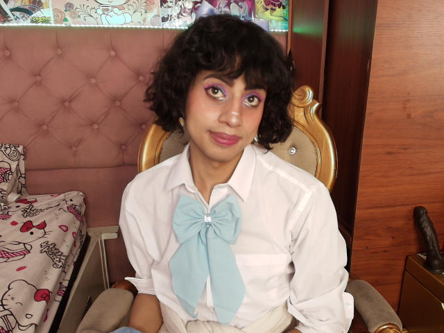 EimyFlorian with curly black hair, pink eyelid makeup, white shirt with blue bow, sitting on a gold chair, smiling, pink tufted headboard, wooden wall, and Hello Kitty bed.