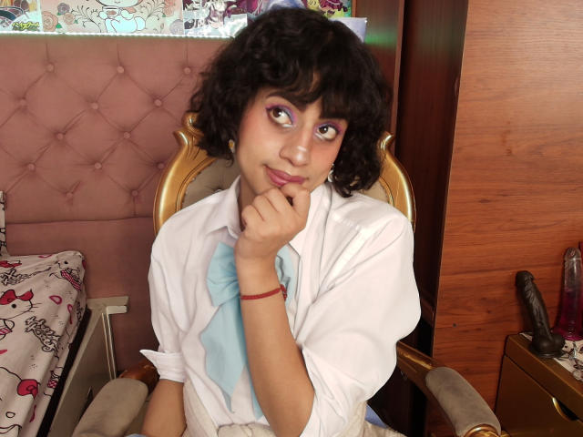EimyFlorian with curly black hair, purple eyeshadow, white shirt, blue tie, hand on chin, smiling, wooden chair, pink tufted headboard, red bracelet, wood wall, bed with Hello Kitty.