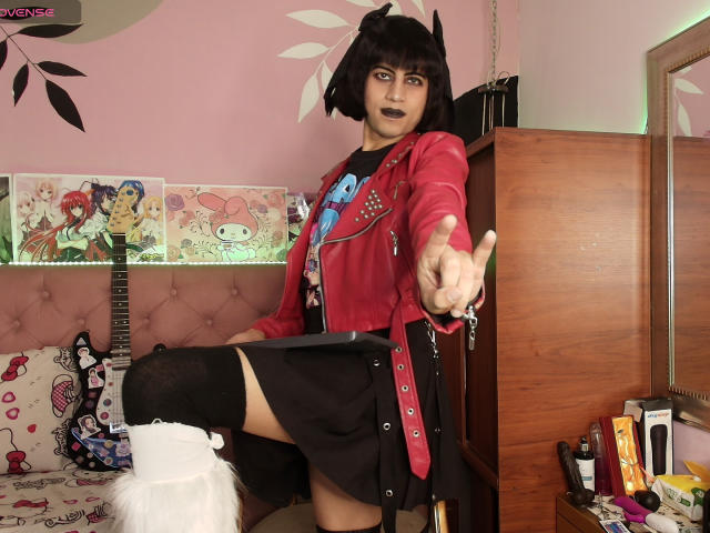 EimyFlorian with black bob, dark lipstick, red jacket, black skirt, thigh-high socks, white furry boot, reaching out, pink bedroom, guitar posters, wooden dresser, mirror, and various items.