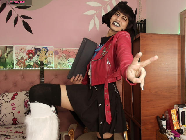 EimyFlorian with black bob, grinning, wearing red jacket, black skirt, and thigh-high socks, pointing forward, holding black book, leg up, in pink room, wooden cabinet, guitar in background.