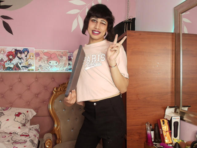 EimyFlorian with short black hair, wearing a pink Paris t-shirt and black pants, holding a knife, winking, and making a peace sign, standing in a pink room.