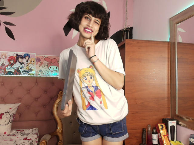 EimyFlorian with curly brown hair, winking, wearing a white "Sailor Moon" tee, denim shorts, holding a large knife, smiling, finger on chin, in a pink room.