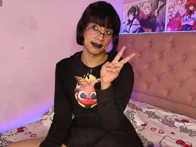 EimyFlorian, with black bob and glasses, dark lipstick, black long-sleeve shirt with cute graphic, sitting on bed, flashing peace sign, purple room lighting.