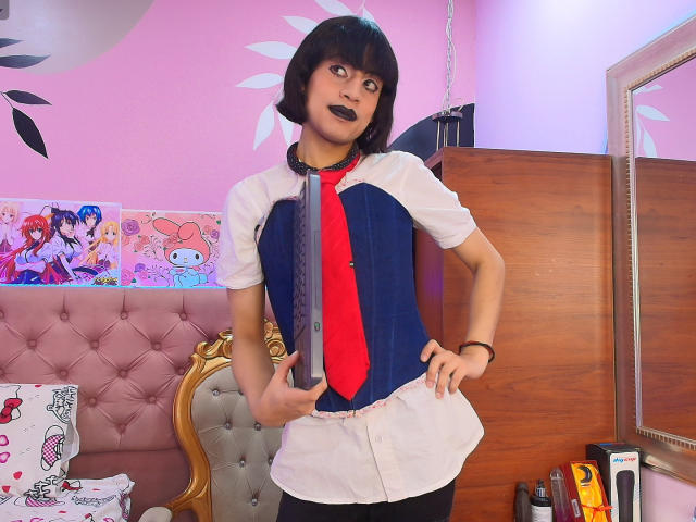 EimyFlorian with a bob haircut, dark lipstick, wearing a white shirt, blue vest, red tie, and black pants, standing in a pink room, hands on hips, looking sideways.