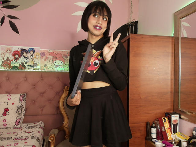 EimyFlorian with a bob, wearing a black hoodie and skirt, holding a ruler, making a peace sign, pink wall, wooden dresser, and anime posters in the background.