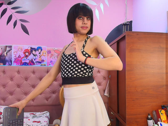 EimyFlorian with a bob, wearing a black polka dot top and white skirt, standing in a room with anime posters, touching her chest, looking at the camera.