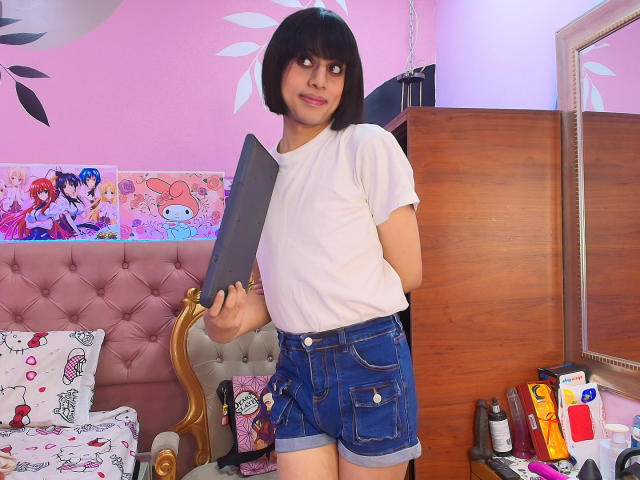 EimyFlorian with short black hair, white t-shirt, and blue denim shorts, holding a large black tablet, standing in a pink room with a wooden dresser and bed.