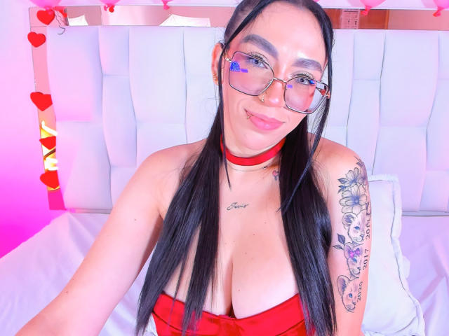 Camille, capelli lunghi e neri, occhiali, collana rossa, top rosso senza spalline, seno grande, braccio tatuato, sorridente, seduta su un letto bianco, luci rosa e rosse..