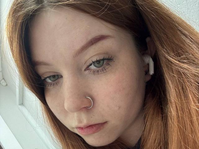LilliamChevalier has red hair, green eyes, a silver nose ring, and white earbuds, with slightly flushed skin and natural makeup, looking softly at the camera.