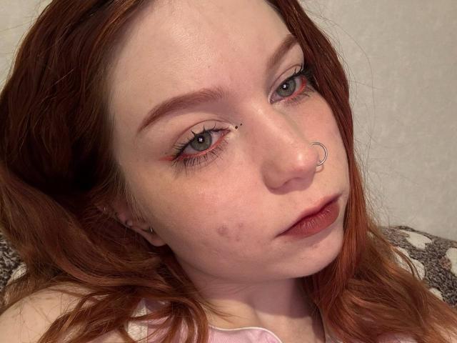LilliamChevalier's face, red-haired, with a nose ring, light freckles, small acne marks, and pink lipstick, staring up with green eyes, eyeliner, and subtle eye makeup.