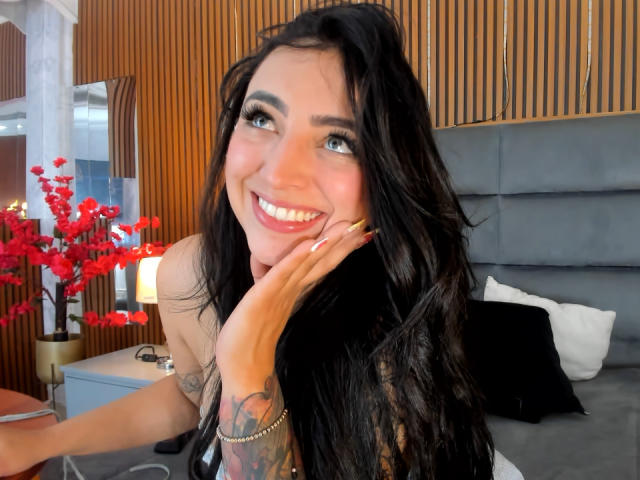 AnastasiaVolkov with long black hair, smiling, hand on cheek, showing off bright red lipstick and tattoos on her right arm, sitting on a bed with red flowers nearby.