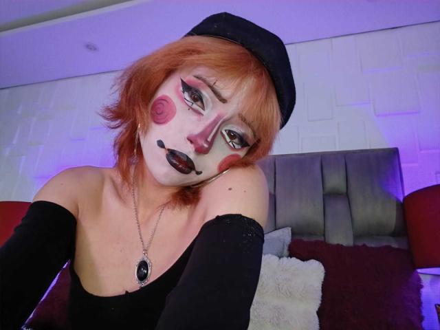 ViolettClay with red hair, black makeup, and a black hat, wearing an off-shoulder black top, showing tits, and a necklace, leaning on a grey headboard.