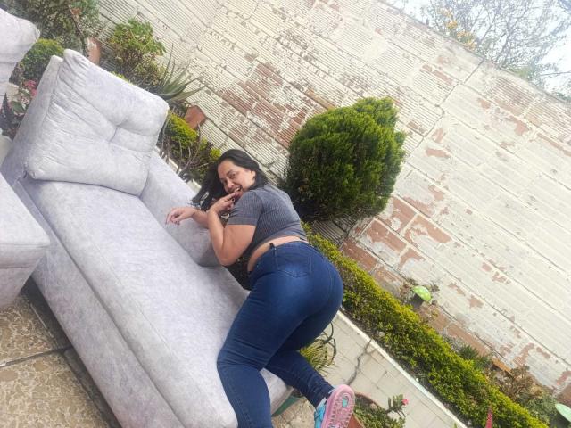KloeDeketAnpenelope, curvilínea com um traseiro grande, vestindo uma cropped cinza e jeans azuis justos, deitada em um sofá cinza claro, olhando para trás com um sorriso divertido..