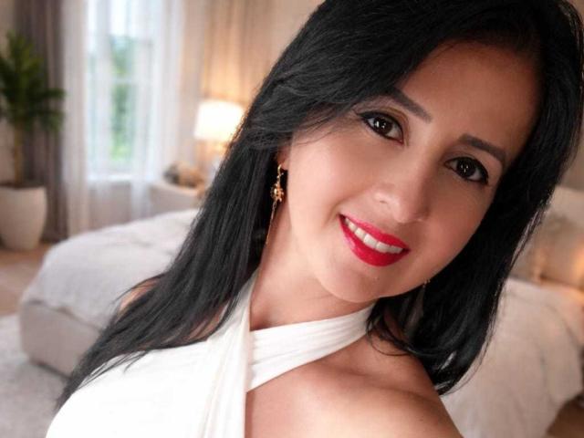 AgathaBrunner has black hair, red lipstick, and a white halter top. She's smiling with brown eyes and gold earrings. Boobs are partially visible. Background is blurred.
