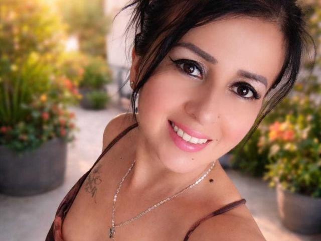 AgathaBrunner has big brown eyes, a cute smile, and small tits. She's wearing a brown top, silver necklace, and has a small black mole on her right cheek.