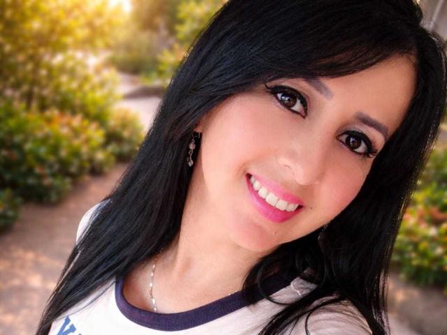 AgathaBrunner has black hair, brown eyes, pink lipstick, and is smiling with white teeth. She's wearing a white shirt with dark sleeves and a silver necklace.