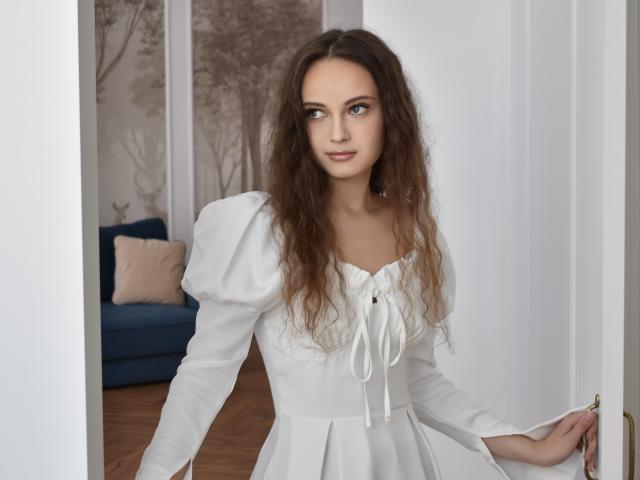 MilenaSparkle has long, wavy brown hair, wearing a white, puffed-sleeve dress with a lace-up front, standing in a doorway with a slight smile, holding the door.