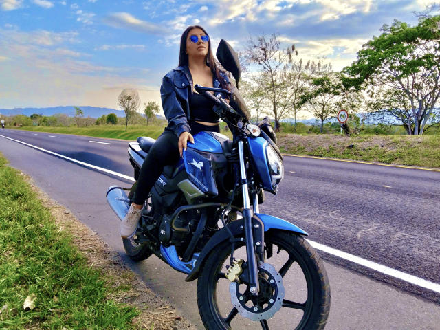 CatalinaRivero with big ass, blue motorcycle, black leather jacket, blue top, black pants, and sunglasses, standing on a road, with a clear blue sky and green trees.