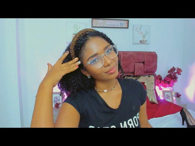 MeganWlliams with curly brown hair, leopard print headband, glasses, black shirt, necklace, hand in hair, medium tits, light brown skin, bedroom with red and pink decor.