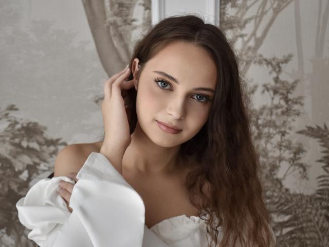 MilenaSparkle with long brown hair, blue eyes, and a slight smile, wearing a white off-shoulder top, hand near ear, looking at camera, soft light on her face.