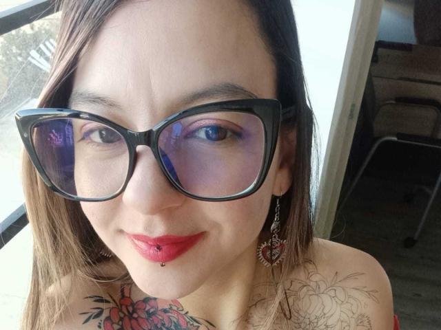 ArianaBlonde with black glasses, red lipstick, and a nose piercing, smiling at the camera. She has tattoos on her chest and a floral earring. ArianaBlonde's tits are not visible.