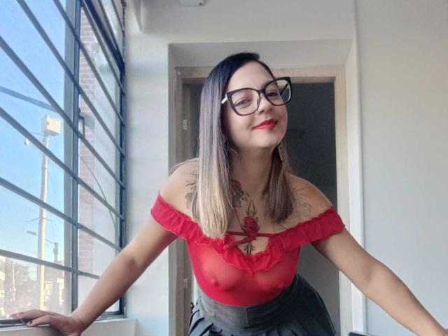 ArianaBlonde with glasses, red off-shoulder top, visible nipples, tattoos on chest, leaning against window, smiling, blonde hair, medium breasts, wearing gray skirt.