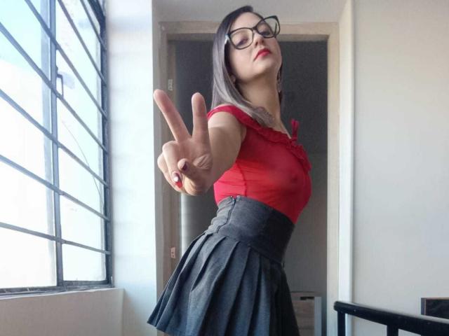 ArianaBlonde with glasses, red top, and black skirt, making a peace sign, showing off her tits, in a room with a window. She's looking up and smiling.