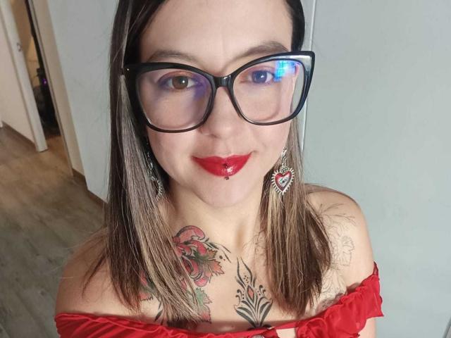 Ariana: Una rubia con gafas, lápiz labial rojo y un piercing en el labio, vistiendo una blusa roja con un hombro descubierto, mostrando tatuajes en el pecho y el cuello, y con aretes, sonriendo a la cámara..