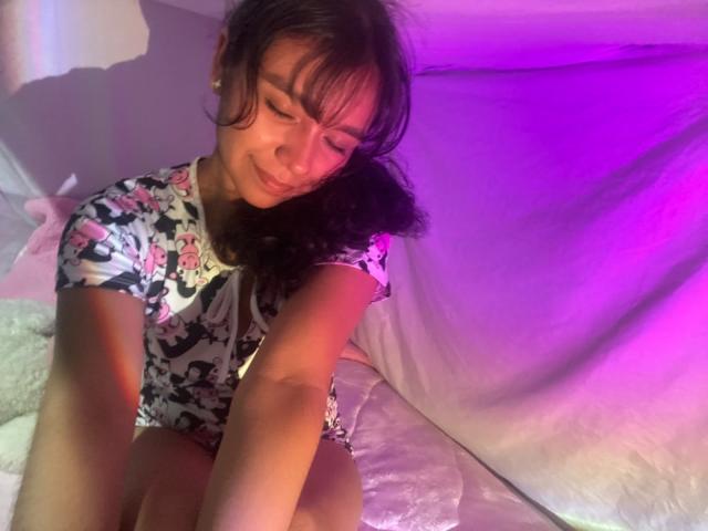 JuliaBennett with curly brown hair, wearing a floral top, sitting on a bed with purple and pink lighting, smiling softly, her arms resting on her knees.