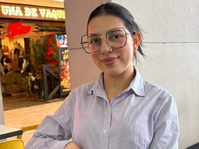Hannah Cruz, usando óculos, camisa roxa de botões, cabelo preto preso em um rabo de cavalo, piercing no nariz e um leve sorriso, com os braços cruzados, sentada em uma mesa amarela..