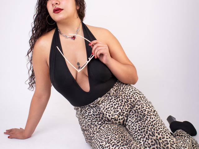 PaulaLoughty with big boobs, wearing a black halter top, leopard pants, holding glasses, red lipstick, curly hair, hoop earrings, and a necklace, sitting on white floor.