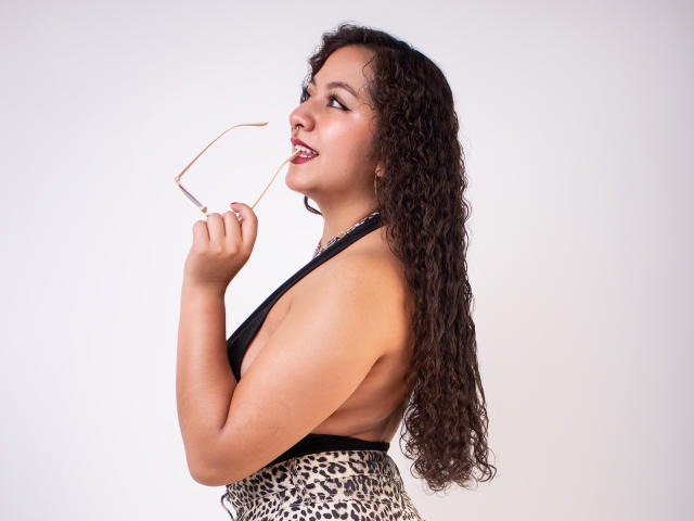 PaulaLoughty with curly brown hair, wearing a leopard-print bottom and black top, holding glasses near her nose, looking sideways with a slight smile, showing her tits.