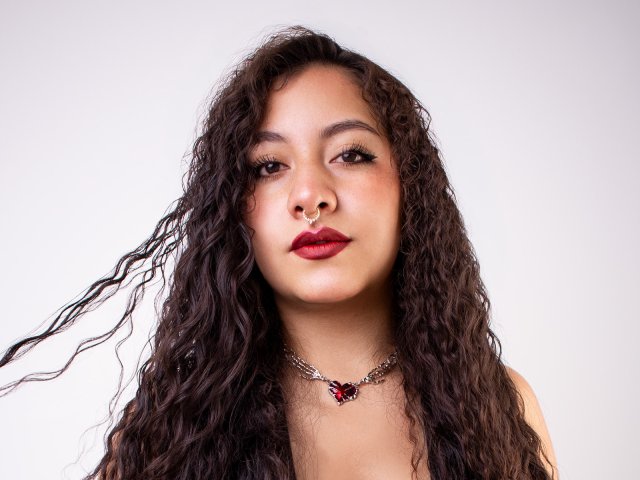 PaulaLoughty with curly dark hair, red lipstick, nose ring, and silver chain necklace with red gem, looking directly at camera.