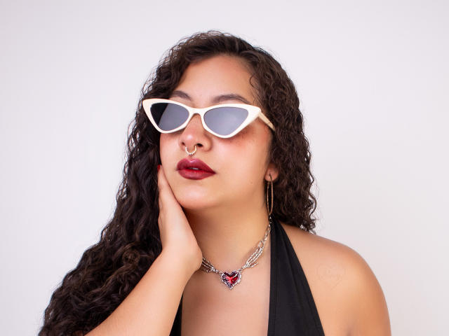 PaulaLoughty with curly dark hair, white cat-eye glasses, nose ring, red lipstick, black halter top, chain necklace with red gem, hand touching cheek.