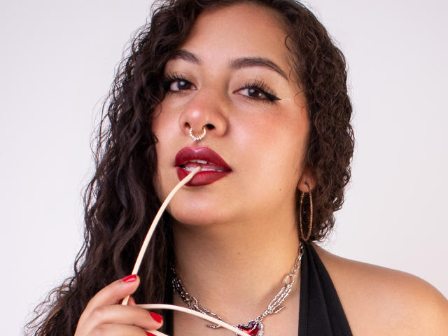 PaulaLoughty with curly dark hair, nose ring, and hoop earrings, sucking on a thin white rod, red lipstick, black halter top, and silver chain necklace.
