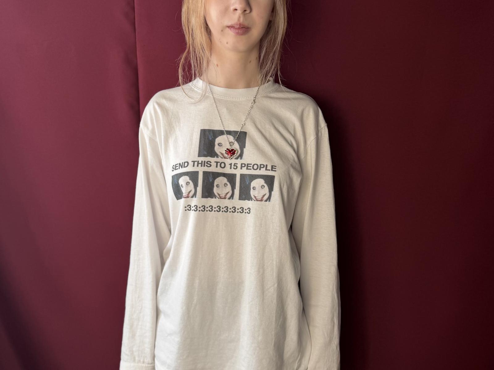 VeniceTototzintle draagt een los wit T-shirt met een foto van de gezichten van vier mensen, en een ketting met een klein rood hart, en staat voor een donkerrode achtergrond..