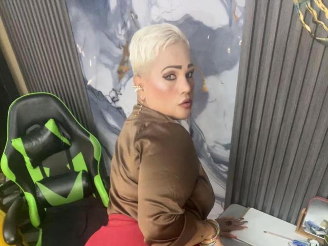 GianellaCox with short platinum blonde hair, brown top, red skirt, sitting on green and black gaming chair, looking back, earrings, subtle makeup, slight smile, medium-sized tits.