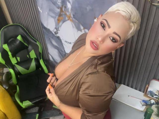 GianellaCox with short blonde hair, brown shirt, deep cleavage, looking up, holding shirt, green and black chair, gold earrings, red lipstick, and a necklace.