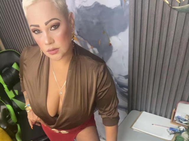 GianellaCox with platinum blonde hair, brown silk top, deep cleavage, red shorts, gold necklace, and earrings, sitting in chair, looking up, sexy, cute, attractive.