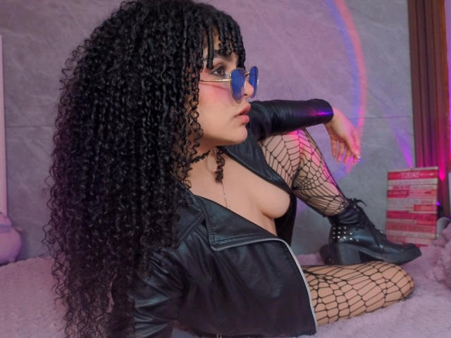 VanessaBlake with curly black hair, glasses, and blue eyeliner, wearing a black leather jacket, fishnet stockings, and black boots, sitting on a pink bed, exposing her tits.