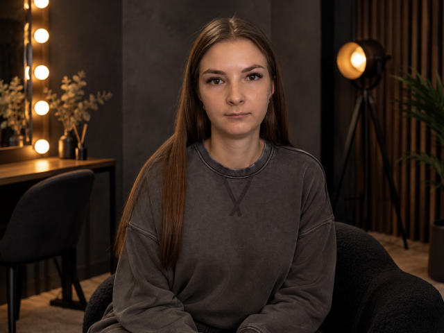 AlisiaTeen heeft lang bruin haar, draagt een grijze sweater, zit met een neutrale uitdrukking en heeft kleine borsten, in een zwak verlichte kamer met een staande lamp en een spiegel..