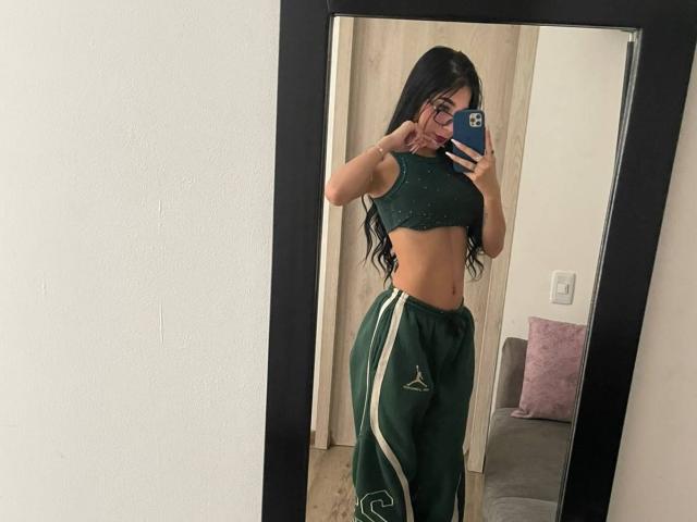 VioletaVelvet with long black hair, glasses, green sports bra, and green pants with white stripes, taking a mirror selfie, showing her toned midriff and curvy ass.